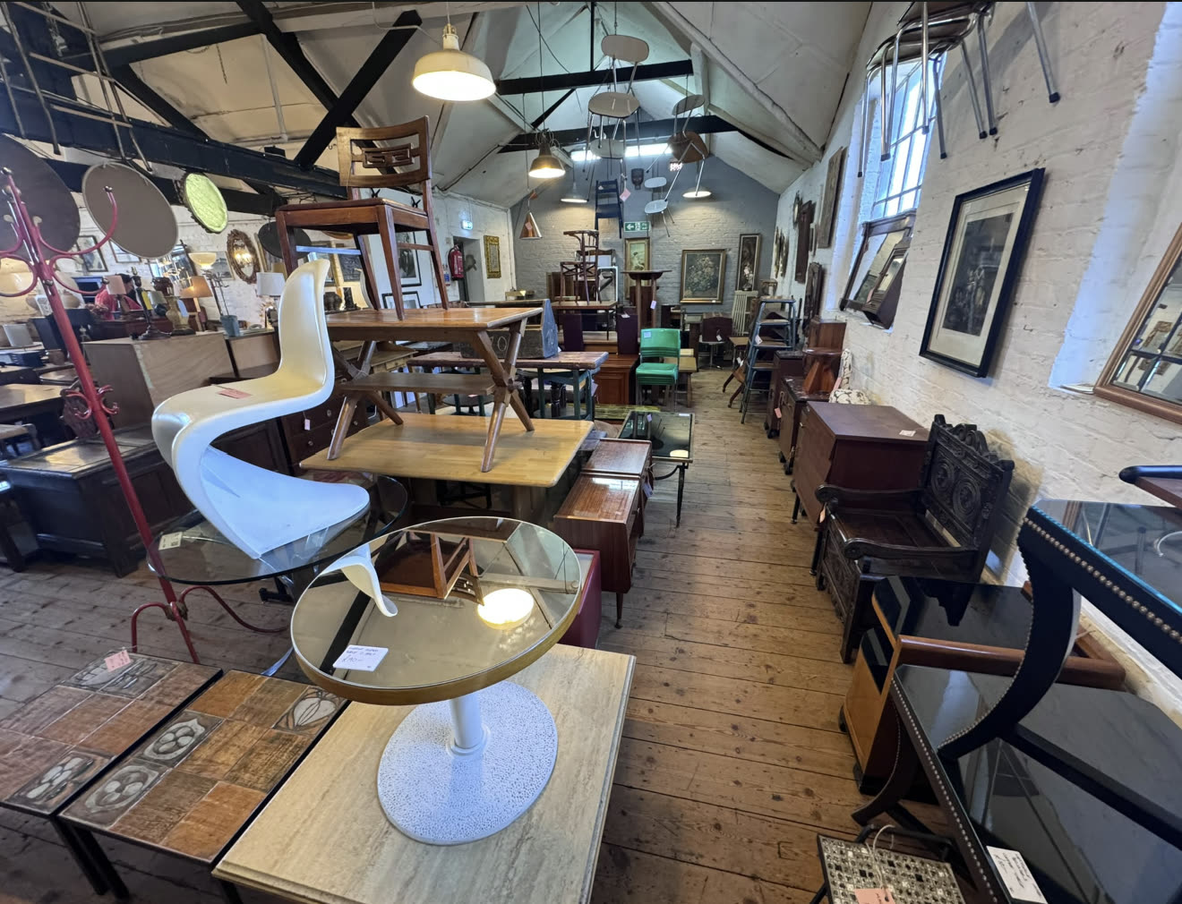 Une longue salle remplie d’antiquités et de vintage : tables, chaises, commodes et miroirs sur un plancher en bois, avec des chaises suspendues aux poutres.